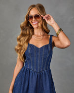 Woman in dark denim corset mini dress with aviator sunglasses, gold bangles, hoop earrings, and layered necklaces