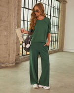 Woman wearing forest green matching lounge set with wide-leg pants and short-sleeve top, holding tumbler and puffer bag with headphones