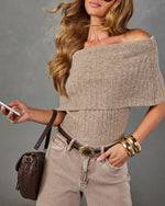 A woman wearing a beige ribbed knit off-the-shoulder top and light beige jeans, accessorized with a brown quilted handbag, gold bangles, and a brown belt, posed against a textured gray studio background.