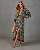A woman models a long-sleeve midi dress with a dark green base and a pink, red, and white floral pattern. The dress features a high slit, a matching fabric belt, and a decorative border at the hem.