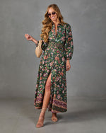 A woman modeling a long-sleeve dark green floral maxi dress with a high thigh slit, patterned hem, and a matching belt, styled with a woven beige clutch and strappy heels against a gray studio backdrop.