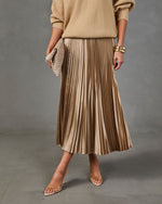 A model wearing a pleated midi skirt in a champagne beige color, paired with a beige knit sweater, a textured clutch, and clear-strap high heels against a gray studio background.