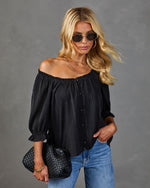 Black off-shoulder blouse with button front and ruffle sleeves styled with blue jeans and woven black clutch bag