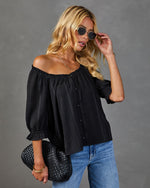 Woman wearing black off-shoulder blouse with button front and puff sleeves, blue jeans, gold sunglasses, and black woven clutch