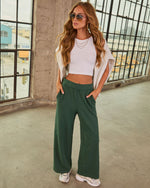 Woman wearing forest green wide-leg pants, white crop top, layered gold necklaces, aviator sunglasses, and chunky sneakers.