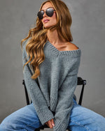 A woman wearing a chunky knit off-the-shoulder sweater in heathered gray, paired with blue jeans and dark sunglasses, seated on a black chair against a textured gray studio background.