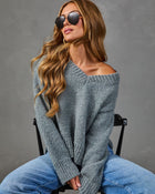 A woman wearing a chunky knit off-the-shoulder sweater in heathered gray, paired with blue jeans and dark sunglasses, seated on a black chair against a textured gray studio background.