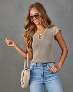 Woman wearing taupe knit cap sleeve keyhole top, high-waist blue jeans, aviator sunglasses, holding cream woven crossbody bag
