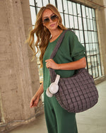 Woman in green athleisure set carrying quilted gray puffer tote bag with white headphones, wearing aviator sunglasses