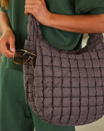 Quilted puffer hobo bag in charcoal gray held by woman in green outfit, with sunglasses in side pocket