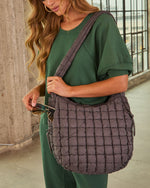 Woman in green outfit wearing quilted charcoal puffer shoulder bag, pulling out sunglasses from side pocket