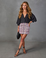 Woman wearing black satin blouse with billowy sleeves, multicolor tweed mini skirt, black strappy heels, and woven clutch