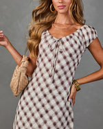 Woman wearing cream and mauve plaid peasant dress with tie neckline, gold bangles, hoop earrings, and woven clutch bag
