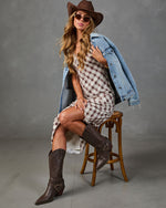Woman in plaid mini dress, denim jacket, brown cowboy hat and knee-high boots seated on wooden stool