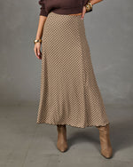 A woman models a long, high-waisted A-line skirt featuring a brown and beige geometric lattice pattern, styled with a dark brown ribbed knit top and brown suede ankle boots against a gray studio backdrop.