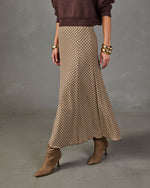 A model wearing a brown and beige patterned knit maxi skirt, styled with a dark brown sweater, gold bracelets, and brown suede ankle boots against a gray studio background.