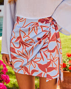 Floral print wrap mini skirt in blue and rust, tied at waist, paired with white long-sleeve top, outdoor setting