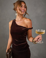 Woman in a brown velvet one-shoulder dress with crystal earrings, gold cuff and fur clutch, holding a champagne coupe