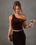 Woman in a brown velvet one-shoulder crop top and matching maxi skirt holding a champagne coupe and faux fur clutch