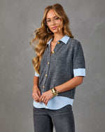Woman wearing charcoal gray short-sleeve button-front sweater layered over light blue collared shirt with gold buttons and gray jeans