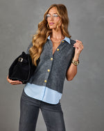Woman wearing gray knit sweater vest with gold buttons over light blue shirt, dark jeans, holding black suede bucket bag