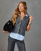 Woman wearing gray knit sweater vest with gold buttons over light blue shirt, dark jeans, holding black suede bucket bag