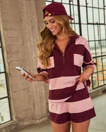 Woman wearing burgundy and pink striped matching shorts set with quarter-zip pullover and coordinating cap, holding smartphone