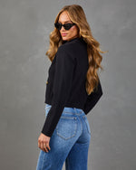 Woman wearing cropped black blazer with gold buttons and blue high-waist jeans, posing with cat-eye sunglasses