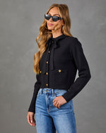 Woman wearing black cropped knit cardigan with gold buttons, bow tie neck, and blue high-rise jeans, styled with cat-eye sunglasses.