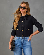 Black cropped cardigan with gold buttons, bow tie neck, worn with blue jeans by blonde model holding clutch