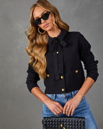 Black knit cardigan with gold buttons, bow tie neck, and front pockets, styled with blue jeans and black quilted clutch