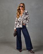 Woman wearing white leopard print oversized shirt jacket, dark wide-leg jeans, brown heeled boots, aviator sunglasses, holding clutch