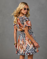Woman modeling abstract ikat-print mini dress with smocked waist, puff sleeves, gold accessories, and round sunglasses