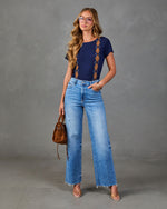 A woman modeling a navy blue short-sleeve top with orange and brown geometric patterns down the sides, paired with high-waisted light blue wide-leg jeans, holding a brown leather handbag against a gray studio background.