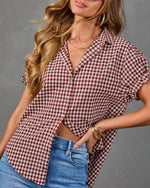 A woman wearing a short-sleeve button-up shirt with a red and brown gingham check pattern, tucked into blue jeans against a gray studio background.