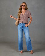 A woman modeling a brown and white gingham short-sleeve button-up shirt paired with high-waisted, wide-leg blue denim jeans, styled with a beige woven clutch and sunglasses against a textured gray studio backdrop.