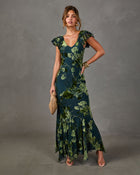 Woman in a dark green floral maxi dress with flutter sleeves, V-neckline and flowy hem, styled with a woven clutch and gold bracelets