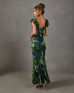 Woman in green floral velvet burnout maxi dress with flutter sleeves, open V-back and handkerchief hem, shown from behind