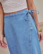 A person wears a textured white crop top and the Maebelle Wrap Tie Denim Maxi Skirt, styled with a bow at the waist. Gold bangles on one wrist complete this chic boho-inspired denim maxi skirt look.