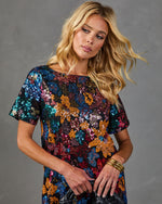Woman wearing multicolor sequin floral short-sleeve dress, holding black clutch, gold bangle bracelet, blonde wavy hair