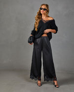 Woman wearing all-black outfit: off-shoulder lace sweater, wide-leg satin lace-trim pants, heels, and woven clutch bag.