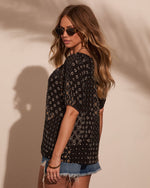 Woman wearing black boho floral print top with short sleeves and denim cutoff shorts, styled with gold hoop earrings and sunglasses