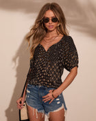 Woman wearing black boho floral print blouse with puff sleeves, distressed denim shorts, sunglasses, and gold accessories