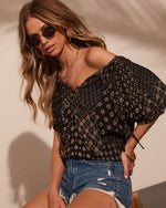 Woman wearing black boho floral print off-shoulder top and distressed denim shorts with gold accessories and sunglasses