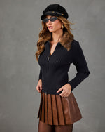 A woman models a black ribbed zip-front sweater with a brown faux-fur collar, paired with a brown pleated mini skirt, black tights, a black newsboy cap, and sunglasses, set against a textured gray studio background.