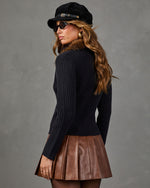 A woman wearing a black ribbed knit sweater, brown pleated faux-leather mini skirt, black hat, and sunglasses, photographed from the back against a textured gray studio background.