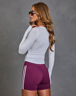 A woman wearing a fitted white long-sleeve athletic top and purple athletic shorts with white side stripes, photographed from the back against a textured gray studio background.