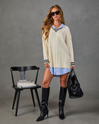 A woman models an oversized cream cable-knit sweater with black and white striped cuffs combined with a light blue and white striped shirt. She is wearing black knee-high leather boots, black sunglasses, and holding a black textured handbag, standing next to a black chair with books on a gray studio background.