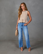 Woman wearing beige peplum knit sleeveless top with rhinestone neckline, wide-leg jeans, clear heels, and woven clutch