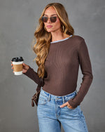 A woman wearing a brown long-sleeve knit top with a light blue crew neck trim, paired with light-wash blue jeans and sunglasses, holding a coffee cup against a gray studio background.